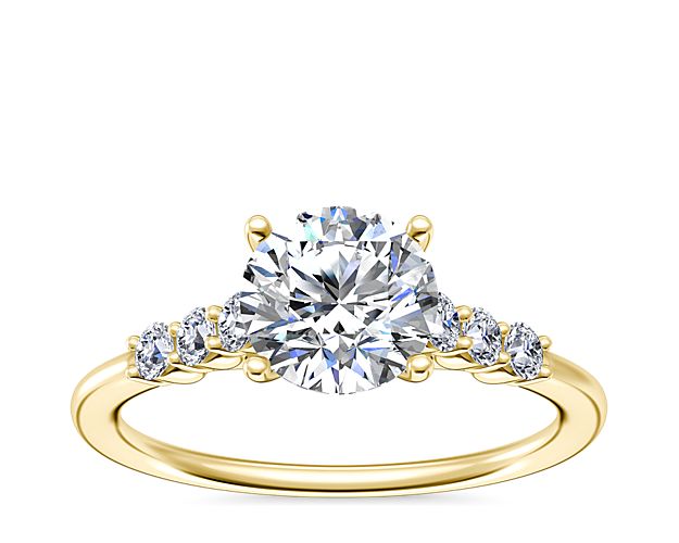 Love shines boldly with this 18k yellow gold engagement ring featuring a brilliant center stone in a sophisticated cathedral setting. Accent diamonds shimmer along either side in shared prong settings to give them bright sparkle.