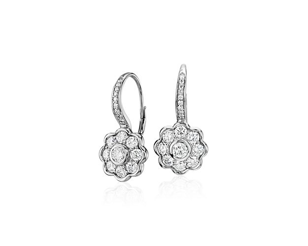 These stunning diamond floral drop earrings in 18k white gold feature a round bezel-set diamond encircled by eight additional diamonds. The stylized floral drops dangles from a pave-set lever-style backing.