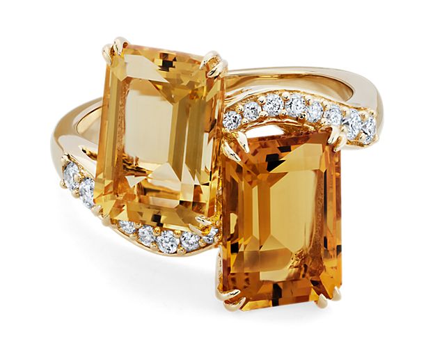 This exquisite yellow gold two-stone ring features two stunning emerald cut citrine gemstones adorned with glistening diamonds.  The citrine gemstones are expertly cut to enhance their bold hue.