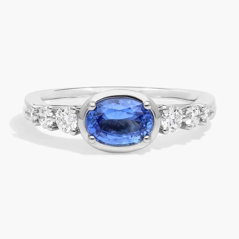 East-West Oval Cut Sapphire Ring With Half Bezel and Diamond Accent In 14K White Gold