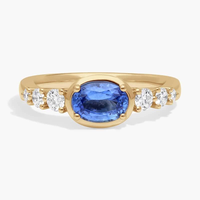 East-West Oval Cut Sapphire Ring With Half Bezel and Diamond Accent In 14K Yellow Gold